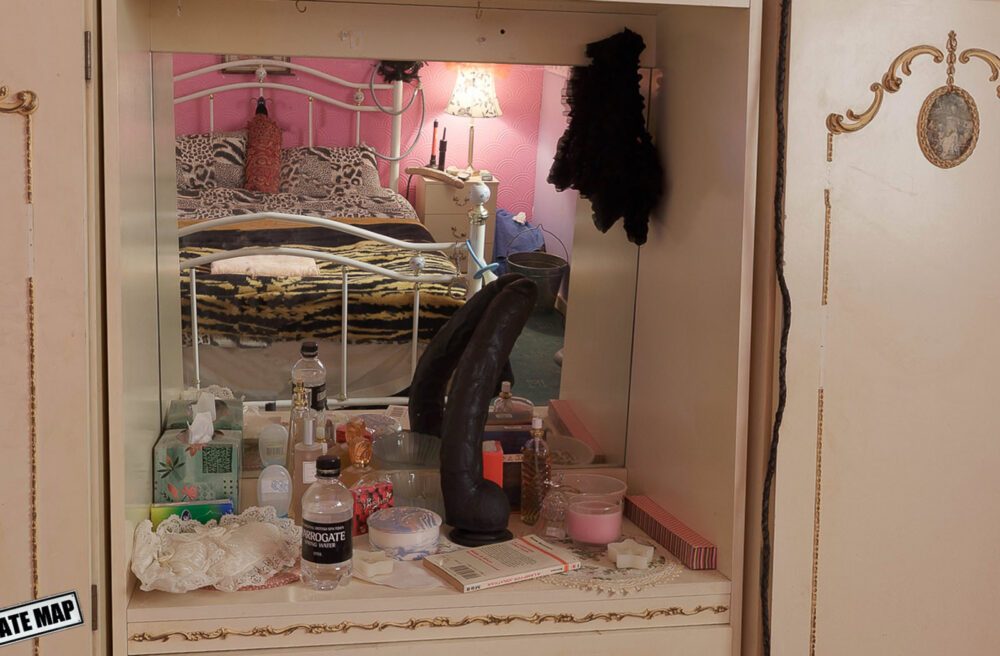 Still image taken from the Shameless virtual tour - view of the dressing table in the bedroom at Lillian's house. The dressing table is cluttered with objects and the bed is visible reflected in the mirror.